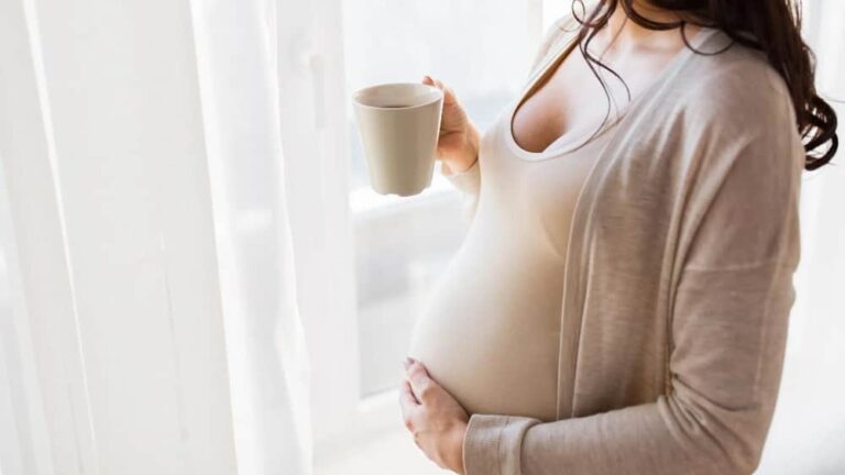 하얀옷을 입은 임신부가 오른손으로 커피잔을 들고 왼손으로 배를 만지고 있는 모습
