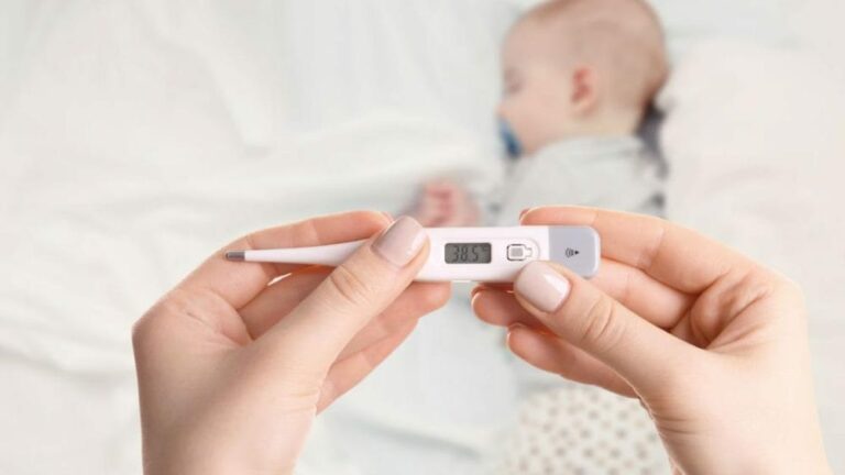 신생아 감기 증상 관련 잠자는 아기 위에서 체온계를 확인하고 있는 손을 포커싱한 이미지