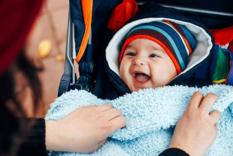 아기 겨울옷 관련 아기가 두꺼운 옷을 입고 웃고 있는데 엄바로 보이는 사람이 이불을 덮어주고 있는 이미지