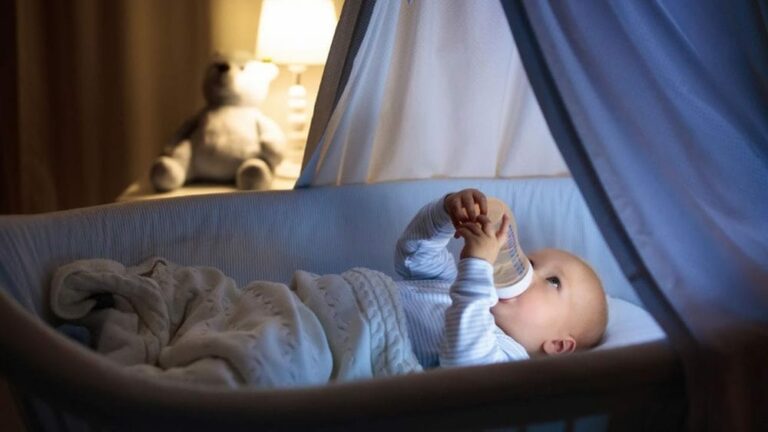 신생아 밤낮 바꾸기 관련 아기가 어두운 방 아기침대에서 양손으로 젖병을 잡고 무언가 마시고 있는 모습