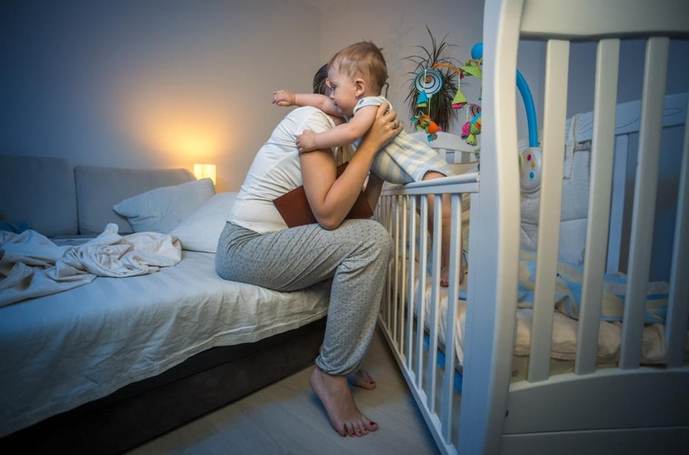 불꺼진 방에서 남자가 앉은 채 아기를 아기침대에서 들어 올려 안으려 하는 이미지