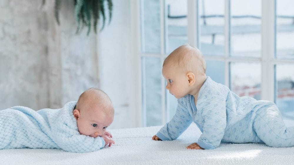하늘색 옷을 입은 두 명의 아기가 서로를 바라보며 나란히 기어 오고 있는 이미지