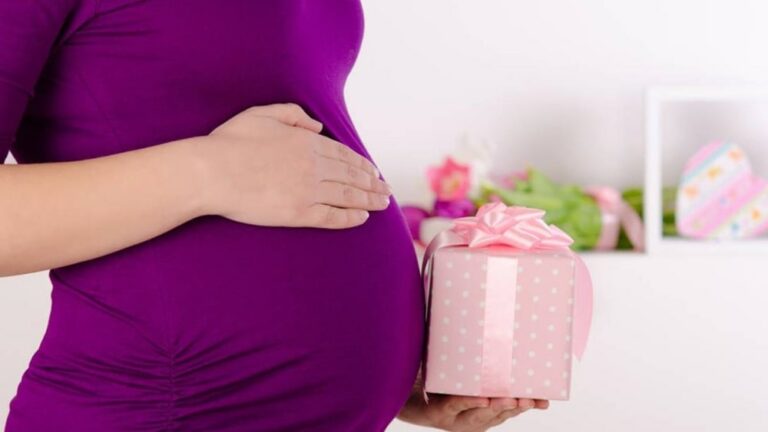 보라색 옷을 입은 임신부가 한손은 배를 만지고 다른 한손으로 선물 상자를 들고 있는 이미지