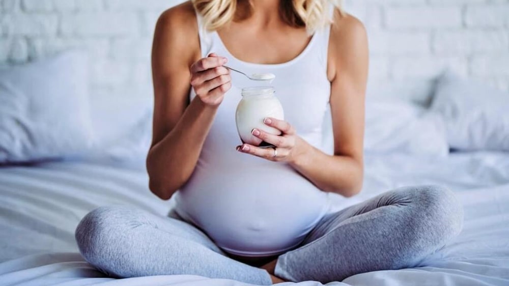 임신부가 양반다리를 하고 앉아서 요거트 병을 들고 약스푼으로 요거트를 뜨고 있는 모습