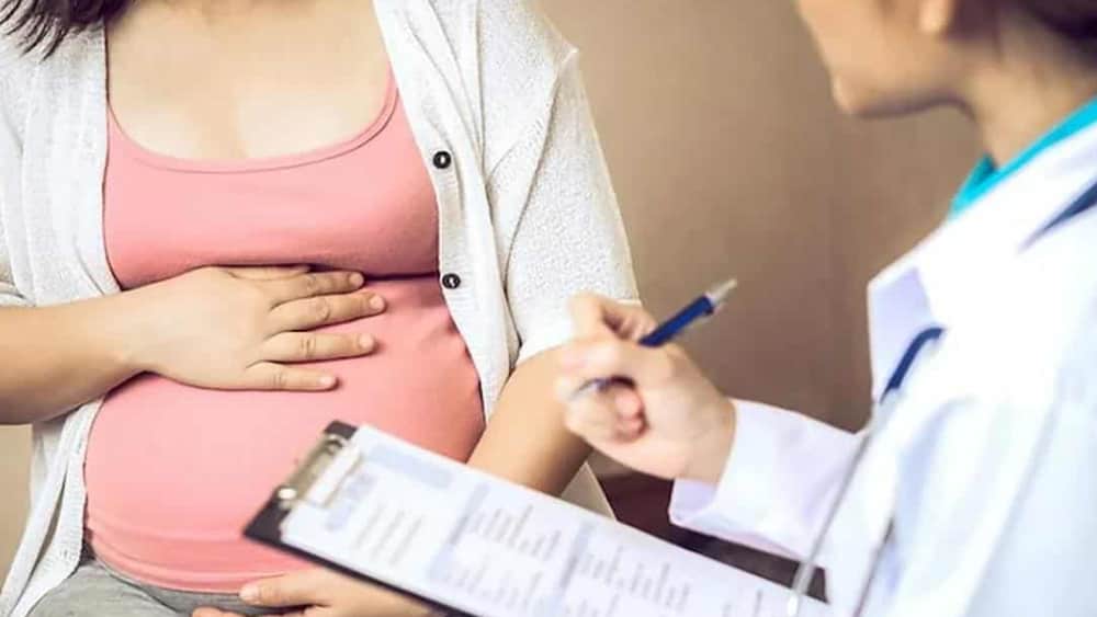 임신부가 앉아서 의사에게 진단을 받고 있고 의사는 서류판에 무언가를 기록하고 있는 이미지