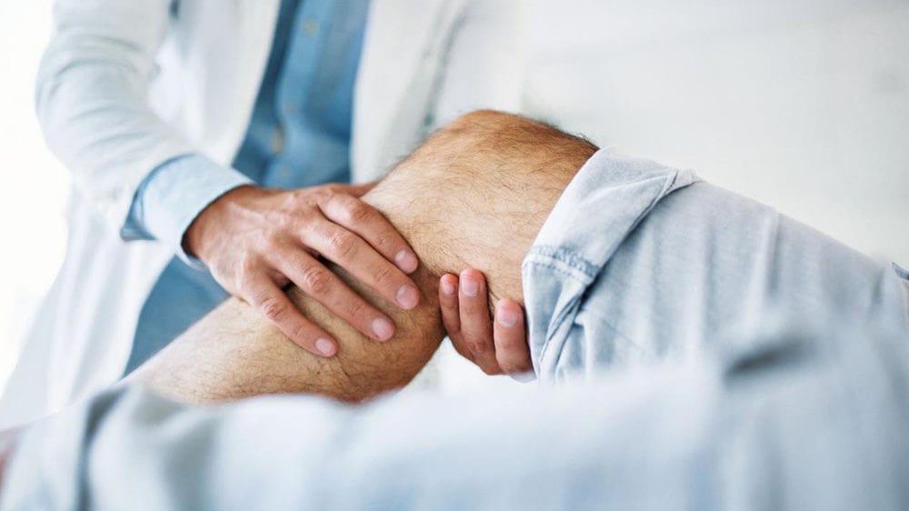 관절 통증 원인 관련 가운을 입은 의사가 환자의 다리 무릎 관절 부분을 손으로 잡고 있는 모습을 포커싱한 이미지