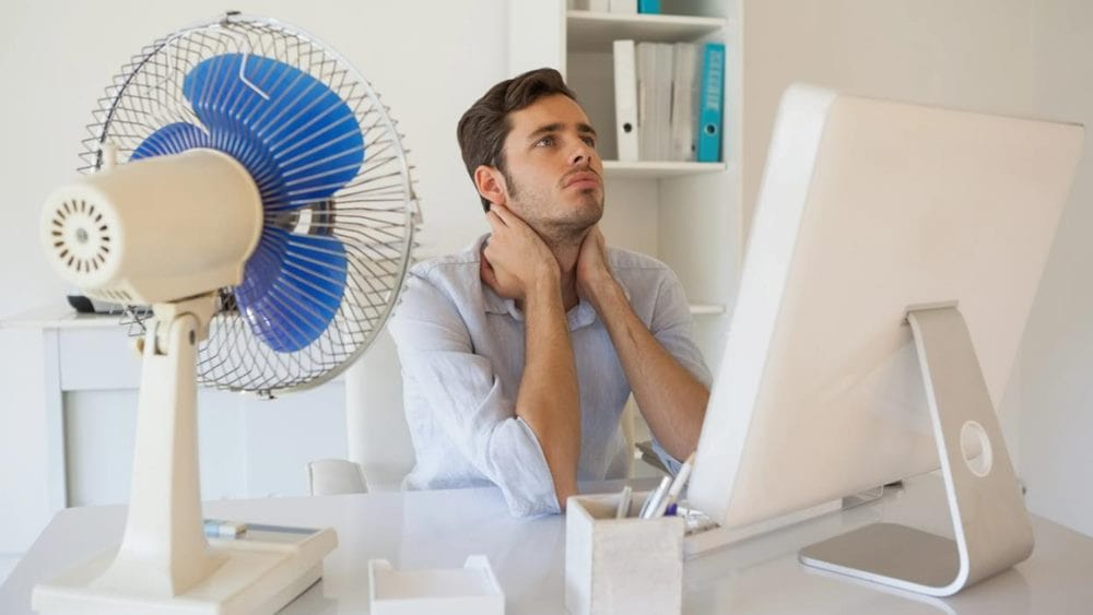 더위 먹는 이유 관련 한 남성이 컴퓨터 앞에 앉아서 양 손으로 목 부분을 괴고 있고 옆에 큰 선풍기가 있는 이미지