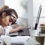 한 여성이 서류가 쌓여 있는 책상 노트북 앞에 엎드려 자고 있는 이미지