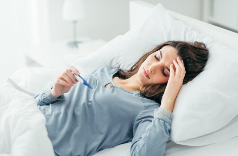 생리전 미열 관련 침대에서 누운 채로 한 손으로는 이마 부분을 짚고 다른 한 손으로 체온계를 보고 있는 여성 모습