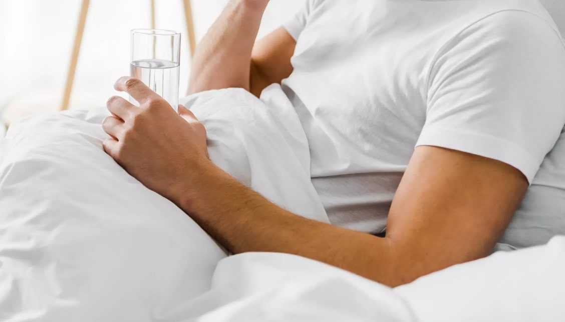 숙취 울렁거림 관련 침대에 기대서 물을 마시고 있는 남성 모습