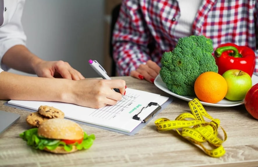 건강한 식단 관련 메모판에 뭔가를 적고 있고 앞에 야채와 줄자, 햄버거 등이 놓여져 있는 모습