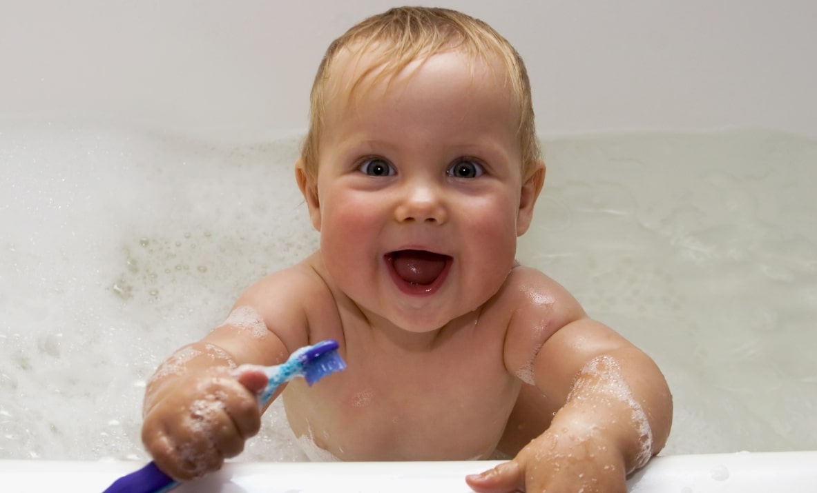 욕조에 들어간 아기가 한 손으로 칫솔을 잡고 웃고 있는 모습