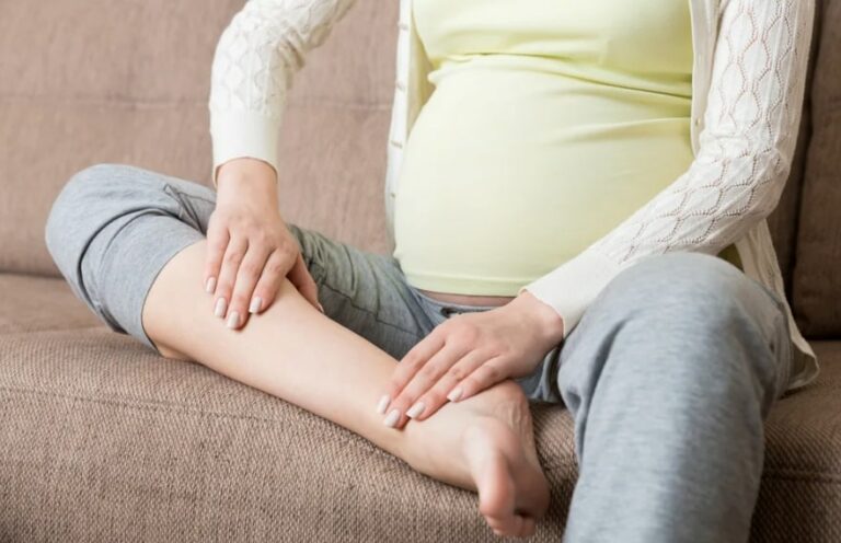 임산부 여성이 소파에 앉아 한쪽 다리를 올리고 양 손으로 주무르고 있는 모습