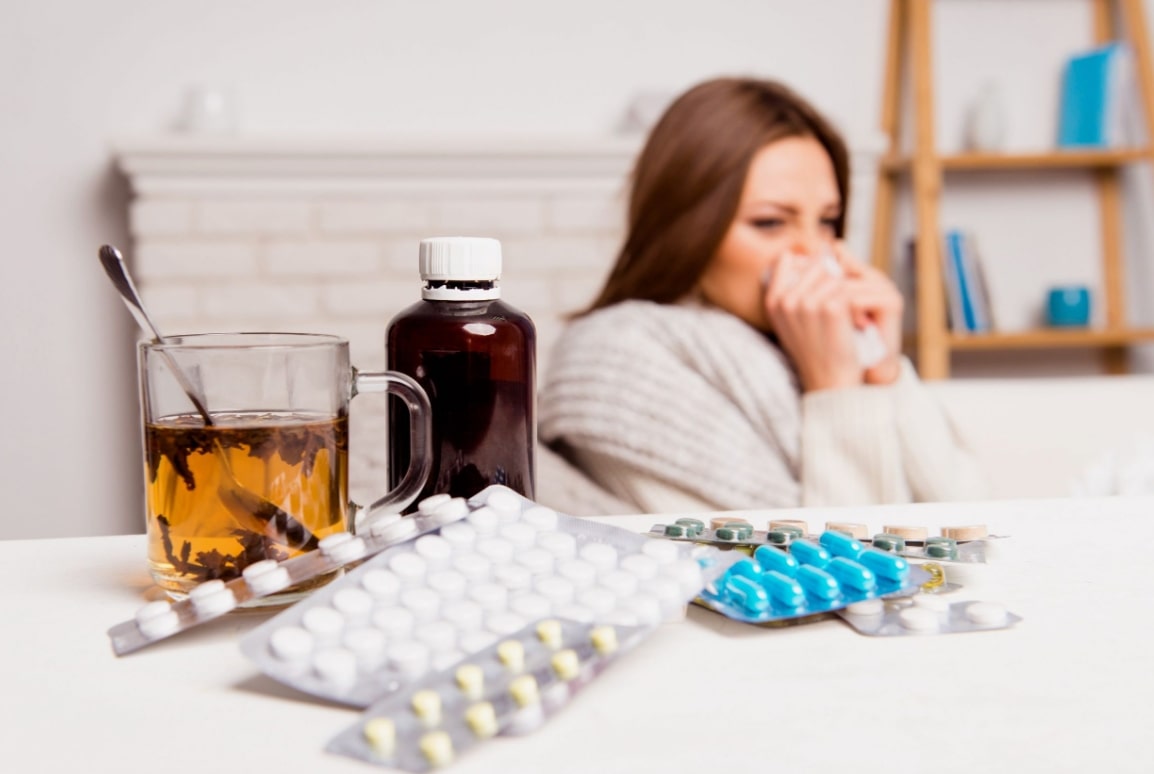 여성이 침대에 누워서 코를 풀고 있고 책상에 약이 많이 놓여 있는 모습